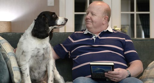 Derek sitting on a sofa looking and patting dog whilst holding a book in other hand.