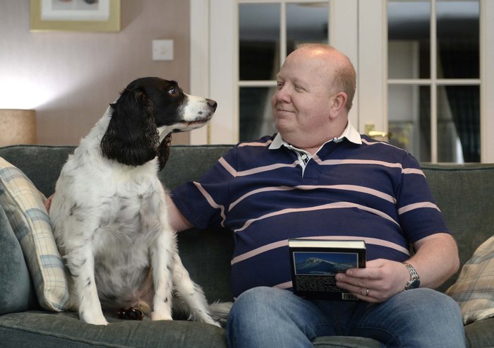 Derek sitting on a sofa looking and patting dog whilst holding a book in other hand.