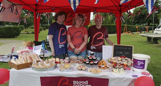 Three women wearing Blood Cancer UK t shirts, smiling behind their beautiful spread of bakes at a bake sale.