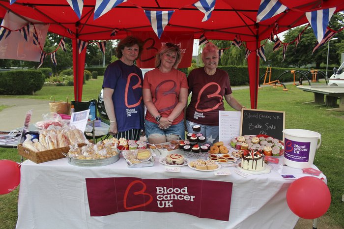 Three women wearing Blood Cancer UK t shirts, smiling behind their beautiful spread of bakes at a bake sale.