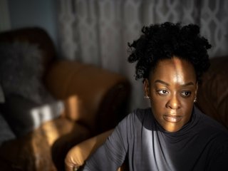 A close up of a black girl standing near a window, the curtains causing light and shadows to create a pattern across her face.