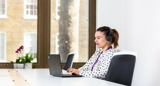 A Blood Cancer UK support line worker on the phone with her headphones on, sitting at her laptop in the office.