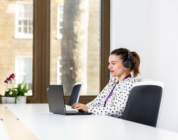 A Blood Cancer UK support line worker on the phone with her headphones on, sitting at her laptop in the office.