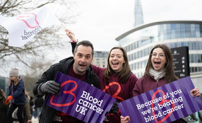 Blood Cancer UK staff members cheering and smiling at an event, holding flags and banners.