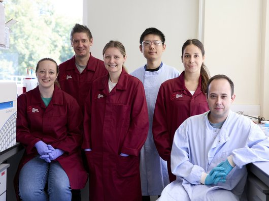 A group of researchers all smiling, wearing Blood Cancer UK lab coats, standing together in the lab.