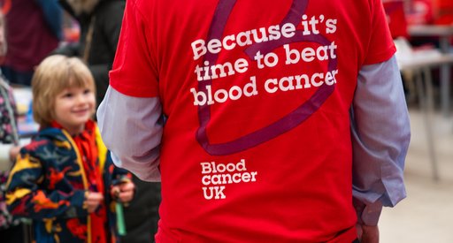 A scene in a village hall. A man stands with his back to us. He has a red Blood Cancer UK t-shirt on that has "Because it's time to beat blood cancer" on the back. There are people in the background and a young boy in a dinosaur coat is smiling up at him.