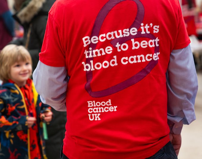 A scene in a village hall. A man stands with his back to us. He has a red Blood Cancer UK t-shirt on that has "Because it's time to beat blood cancer" on the back. There are people in the background and a young boy in a dinosaur coat is smiling up at him.