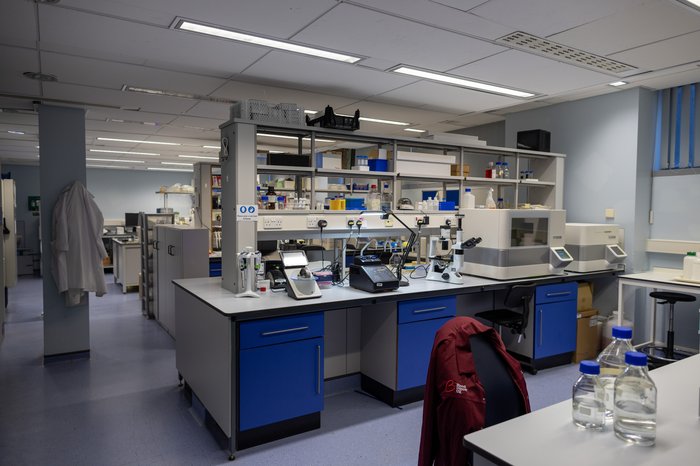 Inside a research lab, work bench with lab equipment and chair with a red Blood Cancer UK lab coat hung over the back.