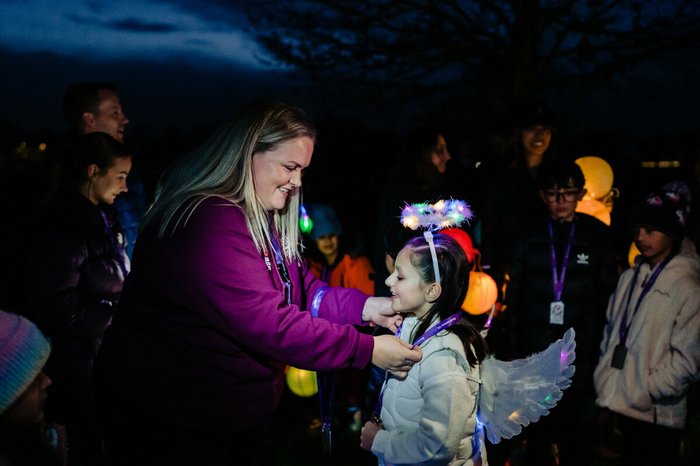 A young girl, Mia, taking part in Walk of Light. A member of Blood Cancer UK is placing a decoration around her neck, and Mia is smiling amidst the fairy lights and night sky.