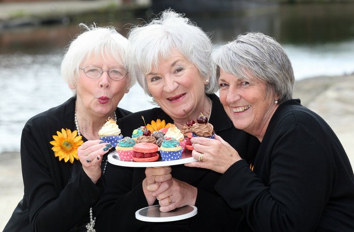 Three of the calendar girls smiling, wearing yellow sunflowers and holding a tray of cupcakes.