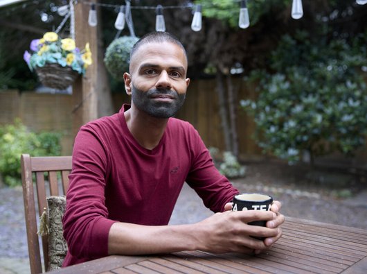 A man is sitting at a garden table, looking at the camera and holding a cup of tea in both hands. He is smiling slightly, and seems thoughtful. There are fairy lights hanging above him and a basket of flowers in the background.