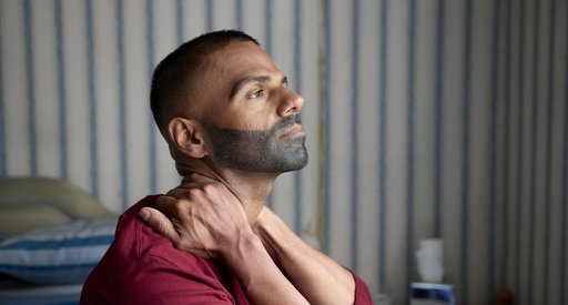 A close up of a man straining his neck by clasping it between his arms and hands. He is looking up slightly, as if towards the sky, with a serious expression.