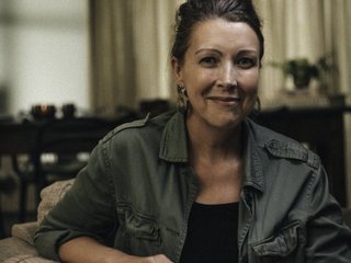 A middle aged woman looks straight at the camera with a slight smile; she is surrounded by a darkly lit room.