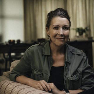 A middle aged woman looks straight at the camera with a slight smile; she is surrounded by a darkly lit room.