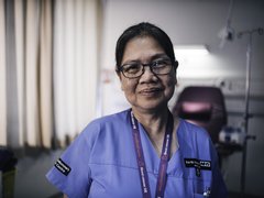 A close up of a healthcare professional wearing blue scrubs and smiling slightly at the camera in a dimly lit room.