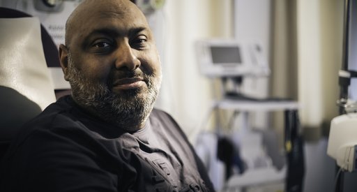 A close up of a man in a hospital bed, looking straight into the camera with a slightly apprehensive smile.