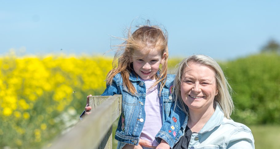 Kirsty holding her daughter up by a fence against a yellow field in the sunshine. They are both smiling with blue sky behind.