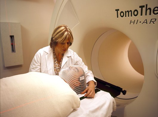 A person lies on a hospital bed in front of a radiotherapy machine with. A clinician stands next to the bed adjusting the person's mesh mask.