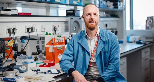 A researcher looking up at the camera, wearing a blue lab coat sitting in a lab, surrounded by wires and scientific equipment.