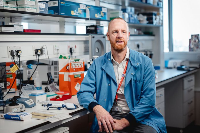 A researcher looking up at the camera, wearing a blue lab coat sitting in a lab, surrounded by wires and scientific equipment.