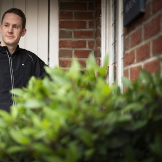 A man stands outside a brick house with a garage door in the background and a plant in the foreground.