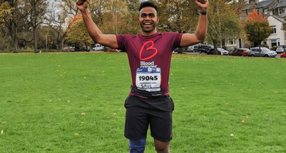 A runner celebrates after a marathon, arms held high in celebration, wearing a Blood Cancer UK T shirt in a park.