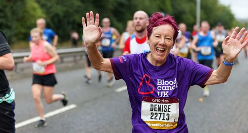 A runner waves to the camera during a marathon; she wears a Blood Cancer UK t shirt.