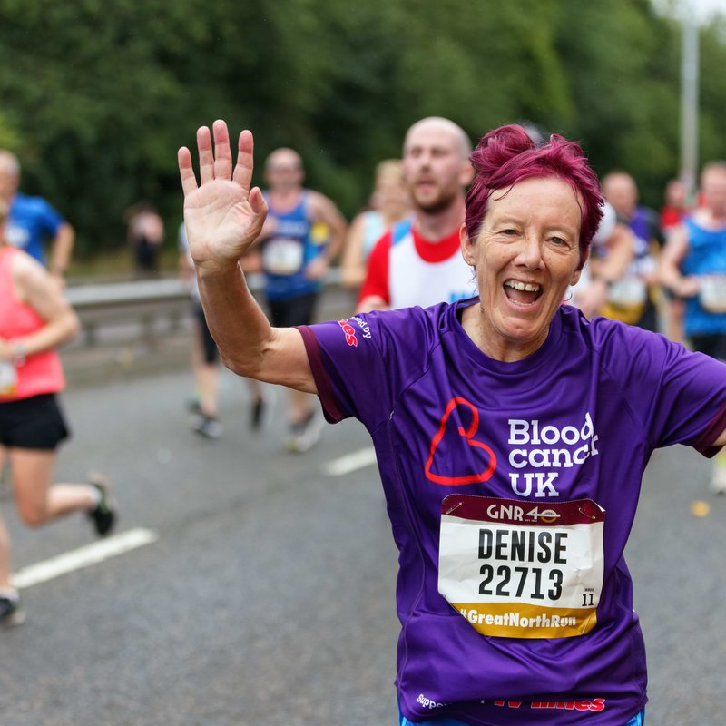 A runner waves to the camera during a marathon; she wears a Blood Cancer UK t shirt.