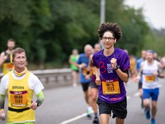 A woman running on a road wearing a purple blood cancer UK t shirt during a marathon.