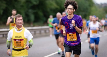 A woman running on a road wearing a purple blood cancer UK t shirt during a marathon.