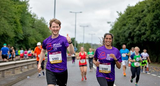 Two runners - a man and woman - smile as they run together wearing Blood Cancer UK t shirts during a marathon.