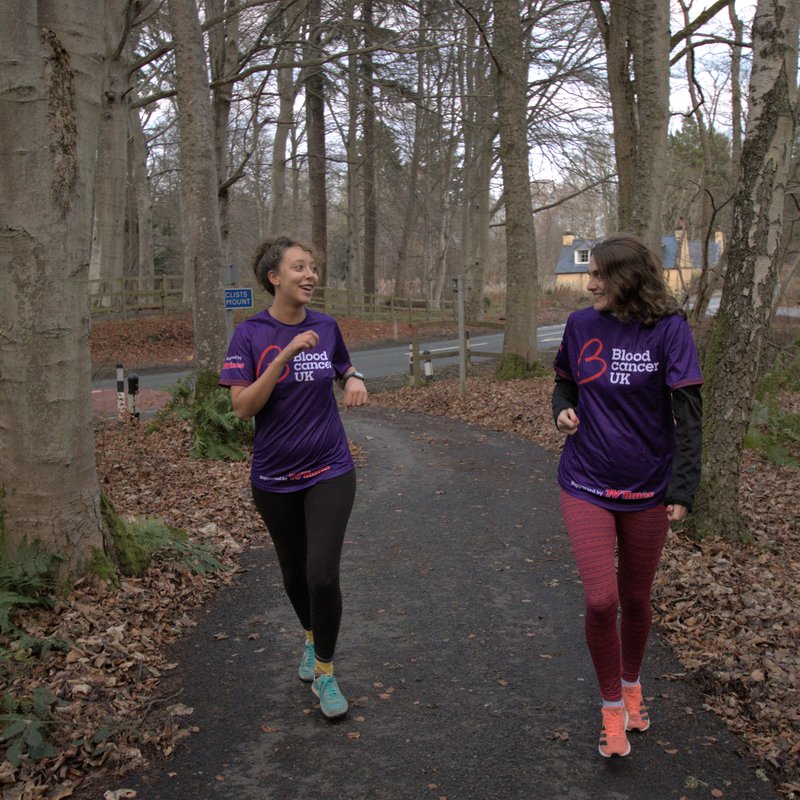 Two female runners running through woodland area looking at each other and smiling in Blood Cancer UK running t-shirts