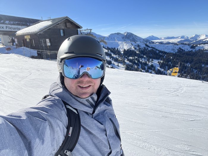 Russell is standing on a snow-covered mountain, smiling at the camera. He is holding a snowboard and wearing ski goggles.