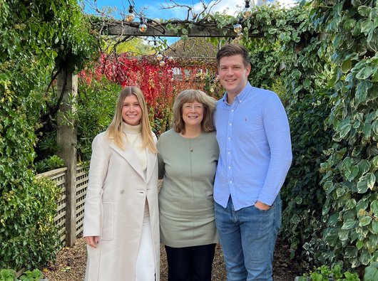 A photo of Samantha Gissel who donates to Blood Cancer UK in memory of her late husband Peter. She is standing in a garden with her children either side of her.