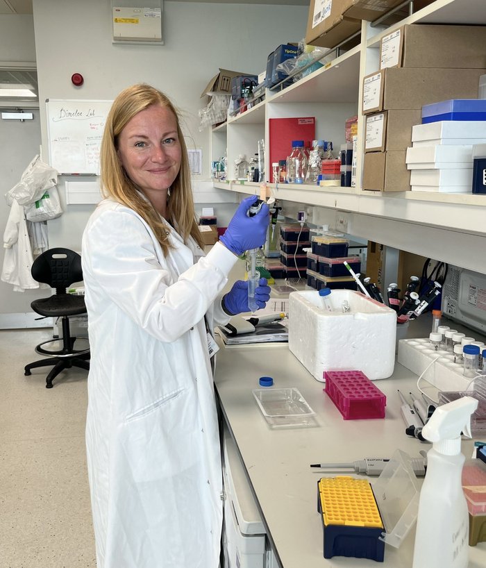 A lady with blonde hair and blue eyes, wearing a lab coat stood in the lab holding a pipette and test tube.