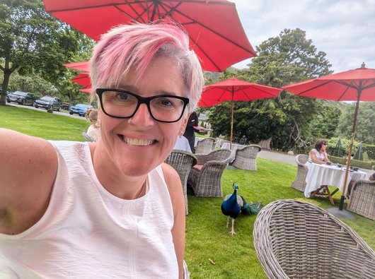 Sarah is sitting outside at a cafe table. She is looking at the camera and smiling, and there is a large peacock walking around next to her.