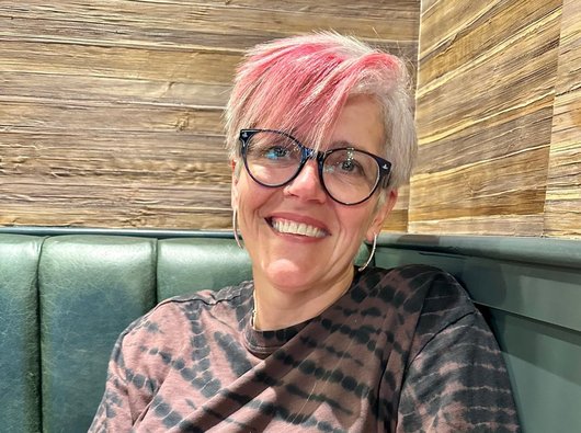 Sarah is sitting on a bench and smiling at the camera. She has short grey hair, and the tips are dyed pink.