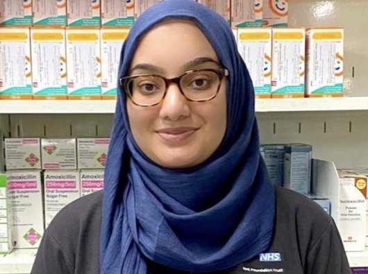 Headshot of Nabeela, who was diagnosed with Hodgkin lymphoma, standing in front of shelves at work which are stacked with various boxes of tablets.