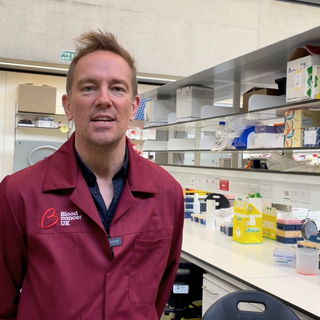 Simon Thomas, stood in the lab wearing a red blood cancer UK lab coat.