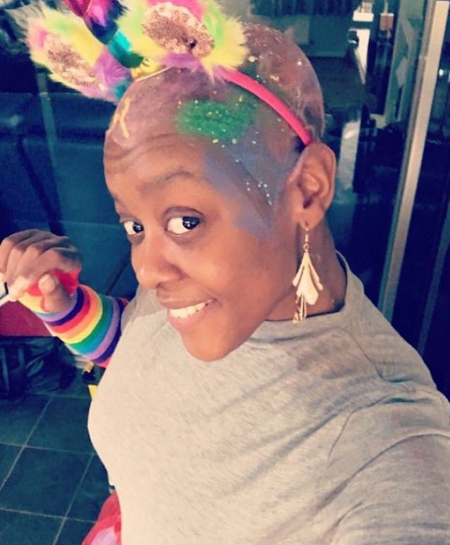 Colourful photograph of Simone with paint and glitter on her bald head, along with a headband with two rainbow coloured ears, and a unicorn's horn. Simone is smiling.
