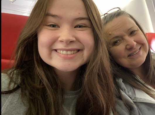 Sophie smiles at the camera. Her head and shoulders are in shot and it looks like she is sat on a train. Her mum is next to her, also smiling at the camera.