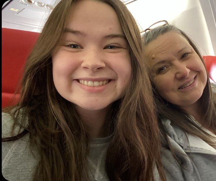 Sophie smiles at the camera. Her head and shoulders are in shot and it looks like she is sat on a train. Her mum is next to her, also smiling at the camera.