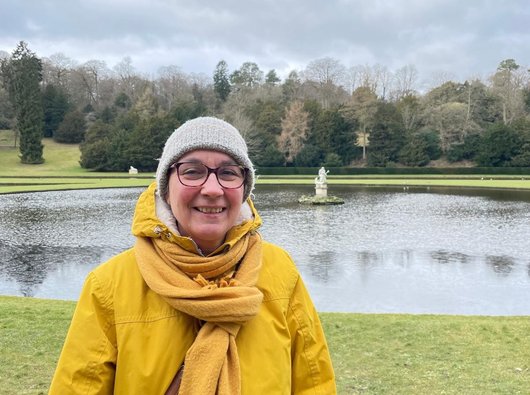 Sue is standing outside on some grass next to a pond. She is looking at the camera and smiling. It looks like it might be raining, and she is wearing a bright yellow coat with a matching yellow hat.