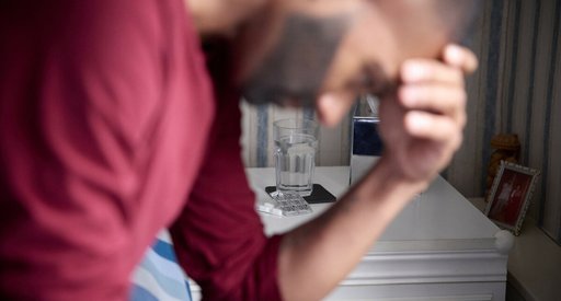 Sunny is sat on the edge of a bed with his elbows on his knees, his head resting in his hands. He is slightly out of focus, and behind him you can clearly see a glass of water and packet of tablets on the bedside table.