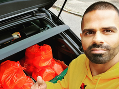 Photo of Sunny standing by his car boot, giving a 'thumbs up', with lots of food shopping in bags visible in his car boot behind him, ready to be delivered around the community.