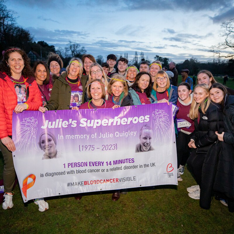A group of a dozen or so people all smiling and holding a banner, taking part in Walk of Light, under an evening sky.