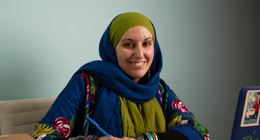 A woman in a headscarf sits at a laptop. She is holding a pen and smiling at the camera.
