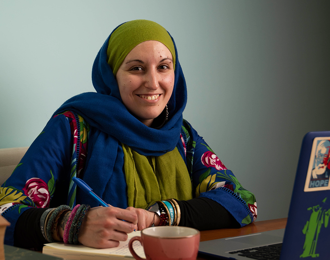 A woman in a headscarf sits at a laptop. She is holding a pen and smiling at the camera.