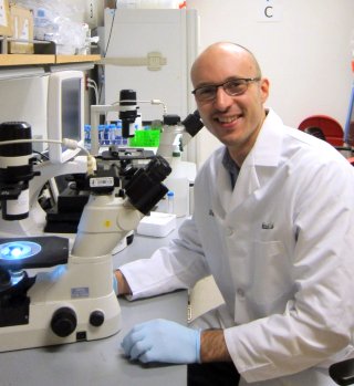 A scientist - Dr Adam Wilkinson - in the lab, smiling at the camera whilst next to a microscope.