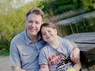 Adrian, who lives with chronic lymphocytic leukaemia (CLL), sits on a picnic table next to a lake, smiling with his child.